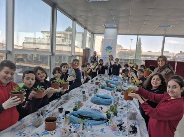 Çocuklar ve öğretmenler, seraya bitki dikimi yaparken mutlu ve öğreniyorlar.